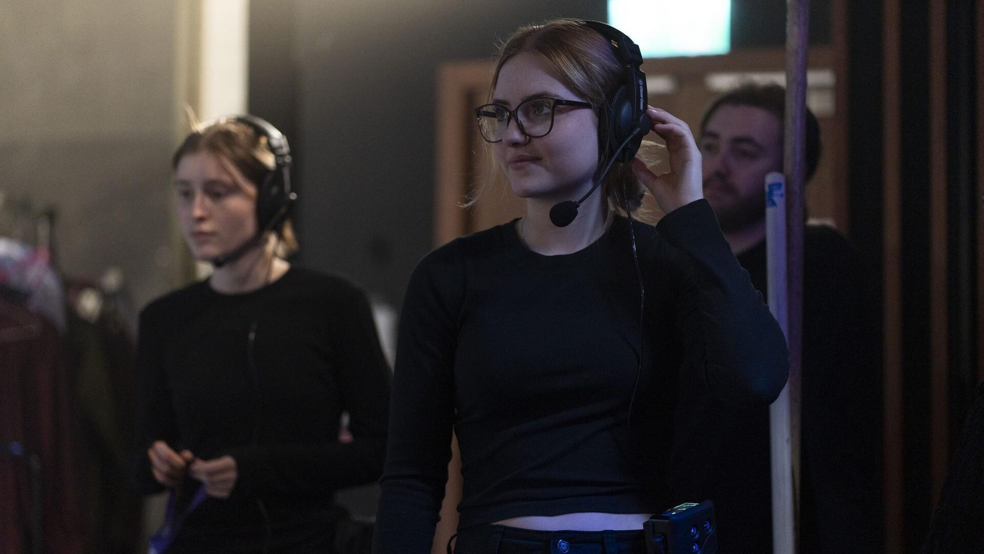 Backstage crew members wearing headset intercoms, one adjusting her headphone while working behind the scenes